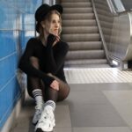 Woman in stylish attire sits by an escalator in a subway station, deep in thought.
