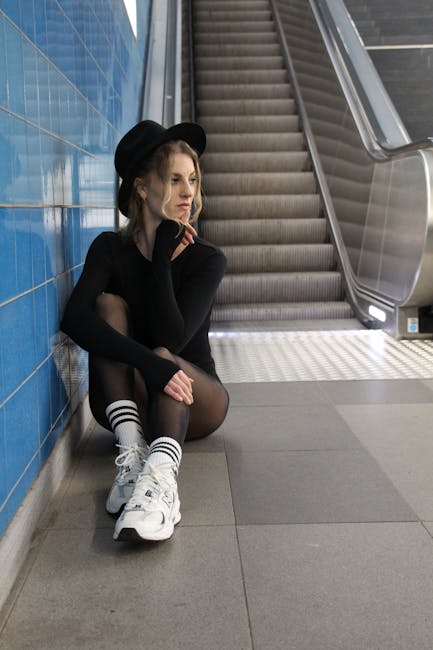 Woman in stylish attire sits by an escalator in a subway station, deep in thought.