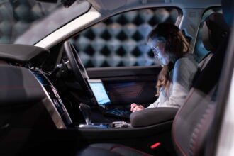 Woman working on a laptop inside a car, testing sound engineering in an isolated chamber.