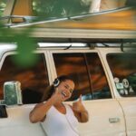 Excited young ethnic female camper showing thumbs up and listening to music in headphones while leaning on car under tent during summer holidays