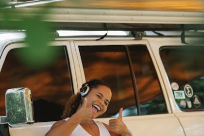 Excited young ethnic female camper showing thumbs up and listening to music in headphones while leaning on car under tent during summer holidays